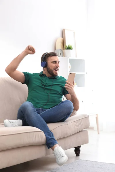 Hombre joven en auriculares con dispositivo móvil disfrutando de la música en el sofá en casa —  Fotos de Stock
