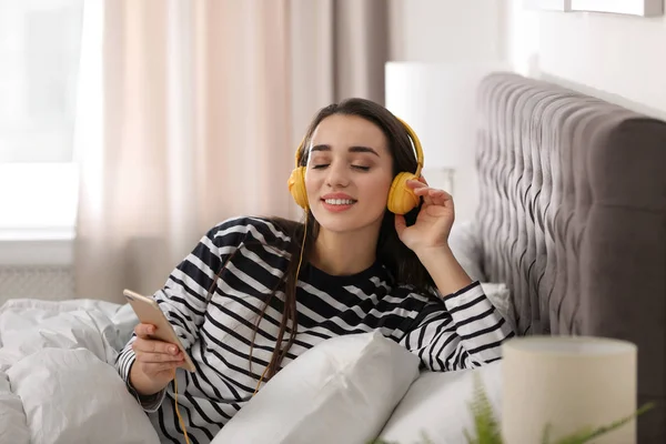 Mujer joven con auriculares y dispositivo móvil disfrutando de la música en la cama — Foto de Stock