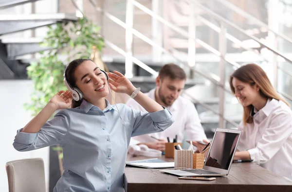 Jovem empresária com fones de ouvido, laptop e seus colegas à mesa no escritório — Fotografia de Stock
