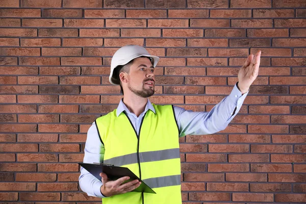 Engenheiro industrial masculino em uniforme com prancheta no fundo da parede de tijolo. Equipamento de segurança — Fotografia de Stock