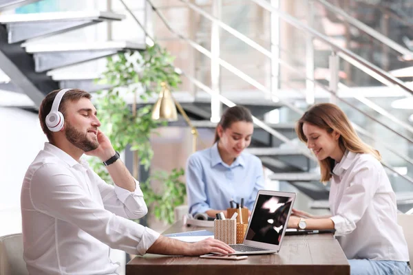 Молодий бізнесмен з навушниками, ноутбуком і його колегами за столом в офісі — стокове фото