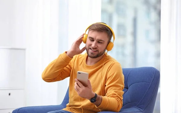 Hombre joven con auriculares y dispositivo móvil sentado en sillón en casa —  Fotos de Stock