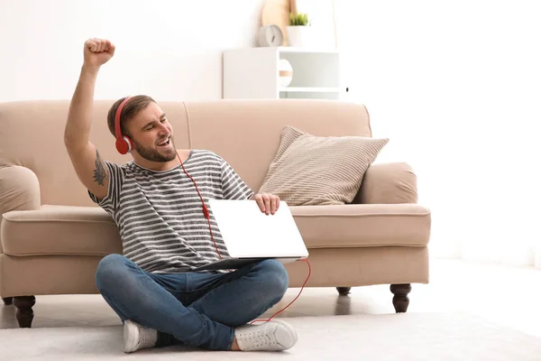 Hombre joven con auriculares y portátil sentado en el suelo en la sala de estar — Foto de Stock