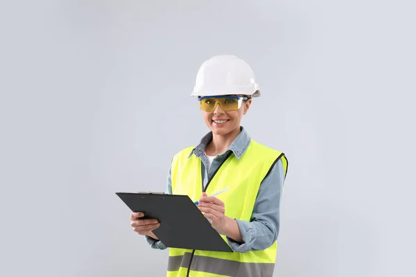 Wirtschaftsingenieurin in Uniform mit Klemmbrett auf hellem Hintergrund. Sicherheitsausrüstung — Stockfoto