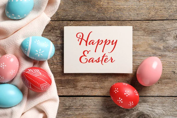 Flat lay composition of painted eggs and card with text Happy Easter on wooden table