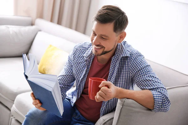 Schöner Mann mit einer Tasse Kaffee beim Lesen eines Buches auf dem heimischen Sofa — Stockfoto