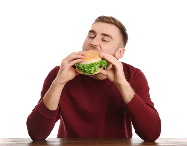 Young man eating tasty burger at table on white background — Stock Photo, Image