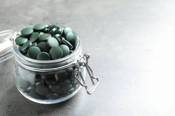 Скляна банка з зеленими таблетками спіруліна на сірому столі, простір для тексту — стокове фото