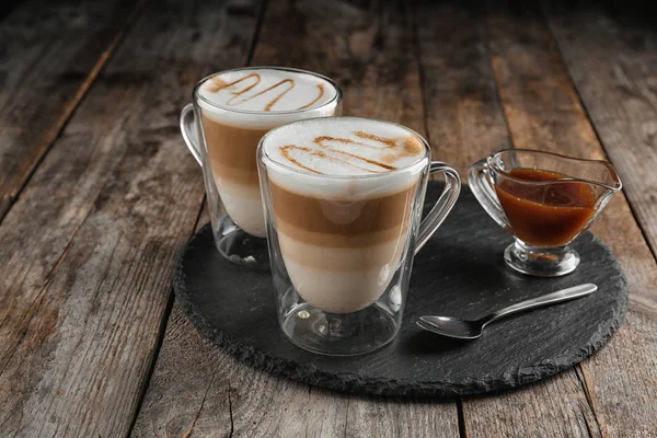 Glass cups of caramel macchiato and gravy boat with syrup served on wooden table — Stock Photo, Image