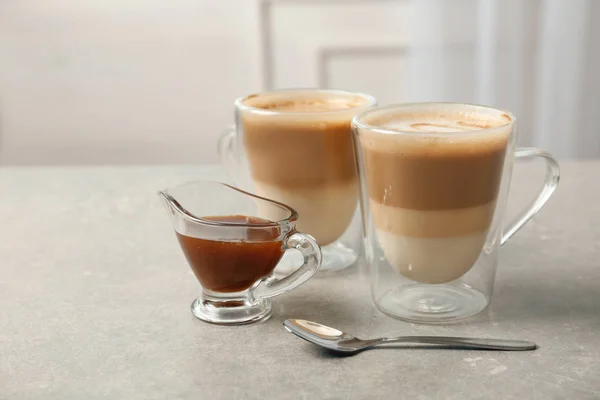 Glass cups of tasty caramel macchiato and gravy boat with syrup on table — Stock Photo, Image