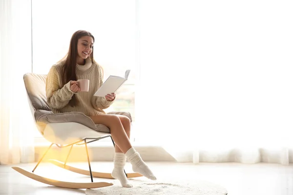 Mujer joven con taza de libro de lectura de café cerca de la ventana en casa, espacio para el texto —  Fotos de Stock