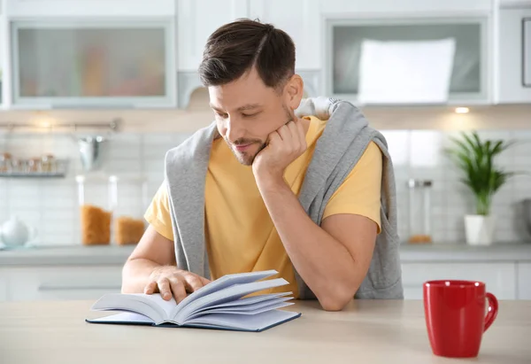 Schöner Mann mit einer Tasse Kaffee und Lesebuch am Tisch in der Küche — Stockfoto