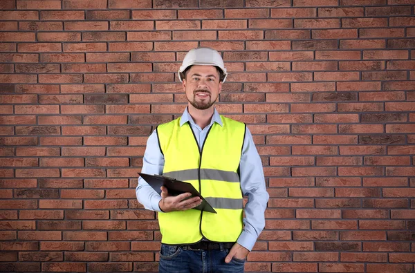 Ingeniero industrial masculino en uniforme con portapapeles sobre fondo de pared de ladrillo. Equipo de seguridad —  Fotos de Stock