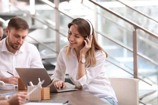 Jonge zakenvrouw met hoofdtelefoons, laptop en haar collega's werkzaam in kantoor — Stockfoto