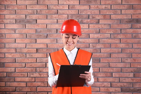 Ingeniera industrial en uniforme con portapapeles sobre fondo de pared de ladrillo. Equipo de seguridad —  Fotos de Stock