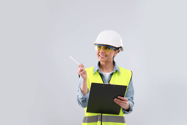Female industrial engineer in uniform with clipboard on light background. Safety equipment