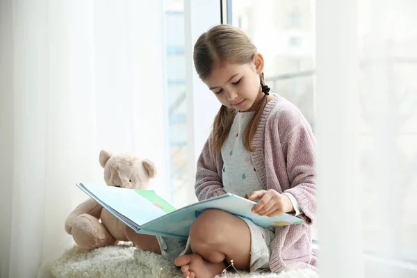Χαριτωμένο κοριτσάκι ανάγνωση βιβλίο κοντά στο παράθυρο στο σπίτι — Φωτογραφία Αρχείου