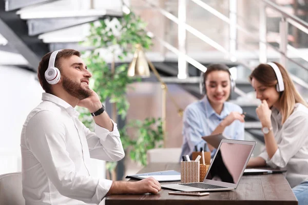 Joven hombre de negocios con auriculares, portátil y sus colegas en la mesa en la oficina —  Fotos de Stock