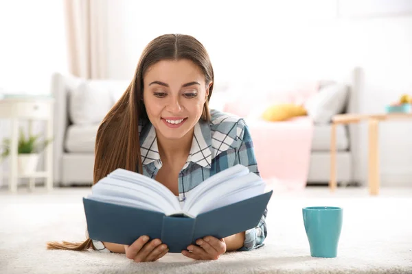 Красива молода жінка з чашкою кави читає книгу на підлозі вдома — стокове фото