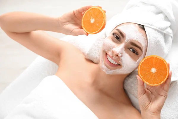 Wanita cantik dengan masker wajah meremajakan memegang irisan jeruk di salon spa, di atas pandangan — Stok Foto
