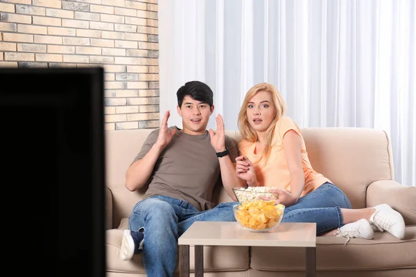 Jovem casal com lanches assistindo TV no sofá em casa — Fotografia de Stock