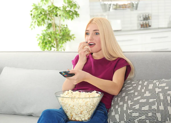 Young woman with remote control and bowl of popcorn watching TV on sofa at home — Stock Photo, Image