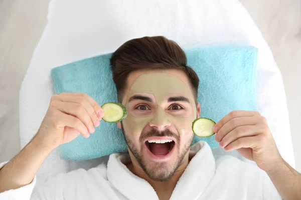 Young man with clay mask on his face holding cucumber slices in spa salon, above view — Stock Photo, Image