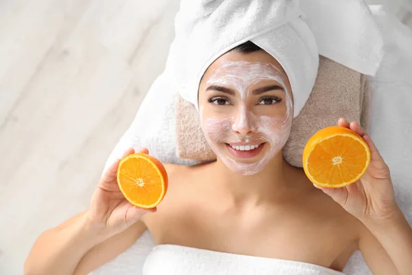 Wanita cantik dengan masker wajah meremajakan memegang irisan jeruk di salon spa, di atas pandangan — Stok Foto