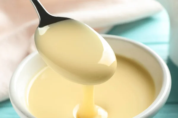 Spoon of pouring condensed milk over bowl on table, closeup. Dairy products — Stock Photo, Image