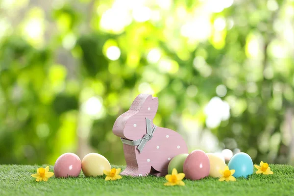 Cute wooden Easter bunny and dyed eggs on green grass against blurred background — Stock Photo, Image