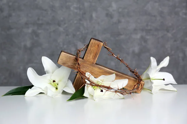 Houten kruis, kroon van doornen en bloesem lelies op tafel tegen een achtergrond met kleur — Stockfoto