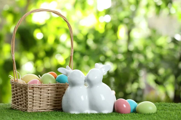 Cute ceramic Easter bunnies with wicker basket and dyed eggs on green grass against blurred background, space for text — Stock Photo, Image