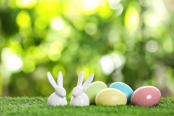 Ceramic Easter bunnies and dyed eggs on green grass against blurred background. Space for text — Stock Photo, Image