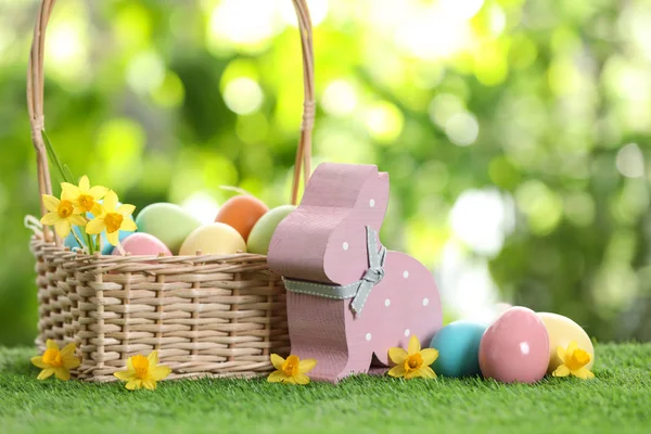 Lindo conejo de Pascua de madera con cesta de mimbre y huevos teñidos sobre hierba verde contra fondo borroso, espacio para texto —  Fotos de Stock