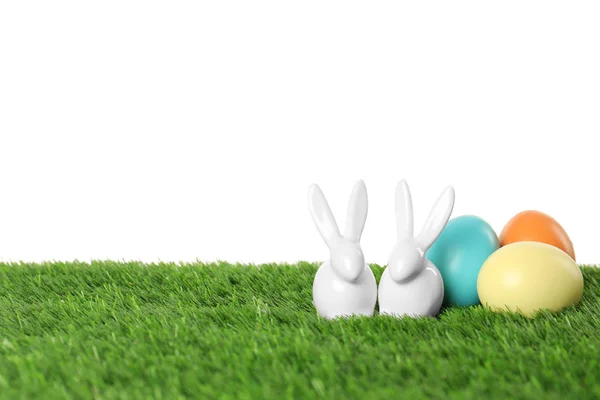 Ceramic Easter bunnies and dyed eggs on green grass against white background, space for text — Stock Photo, Image
