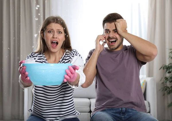 Emotional young woman collecting water leaking from ceiling while her husband calling plumber in living room — Stock Photo, Image