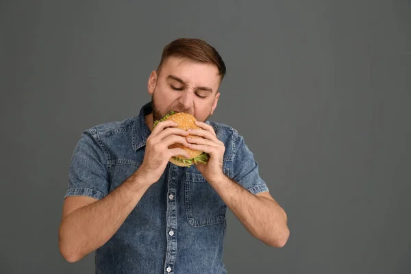 Young man eating tasty burger on grey background. Space for text — Stock Photo, Image