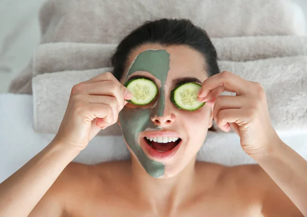 Wanita cantik dengan masker tanah liat di wajahnya memegang irisan mentimun di salon spa, di atas pandangan — Stok Foto