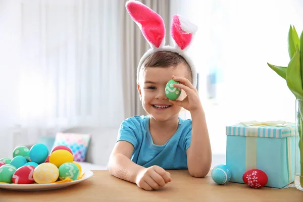 Милый мальчик с повязкой на ушах, играющий с окрашенным пасхальным яйцом в комнате. — стоковое фото