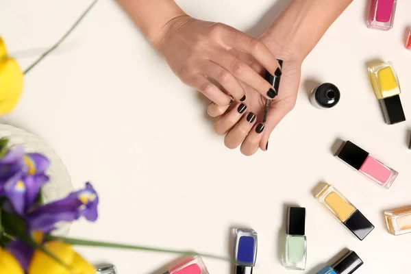 Woman applying nail polish near bottles on white table with flowers, top view — Stock Photo, Image