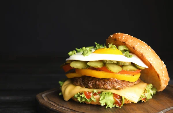 Tasty burger with fried egg on board against dark background, space for text — Stock Photo, Image