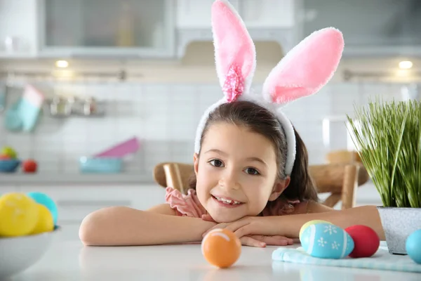 Симпатичная маленькая девочка с повязкой на ушах и раскрашенными пасхальными яйцами, сидящая за столом на кухне, место для текста — стоковое фото