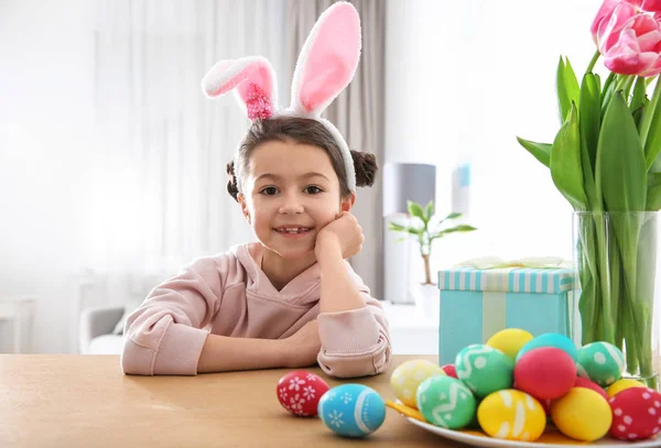 Милая маленькая девочка с повязкой на ушах и раскрашенными пасхальными яйцами, сидящая за столом в комнате — стоковое фото