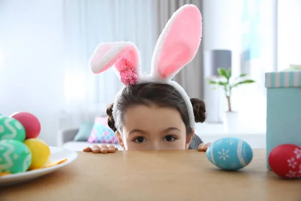 Roztomilá malá dívka, která nosí zajíček uši čelenka schovává za stůl s pomalovaných kraslic v pokoji — Stock fotografie