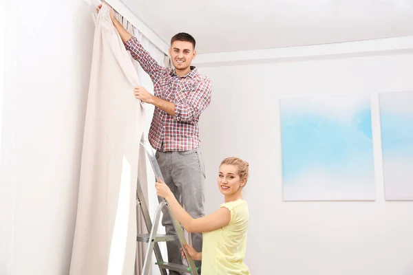 Pareja joven colgando cortina de ventana en el interior. Elemento de decoración interior — Foto de Stock