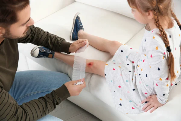 Padre aplicando vendaje en la rodilla lesionada de su hija en casa, primer plano. Primeros auxilios — Foto de Stock