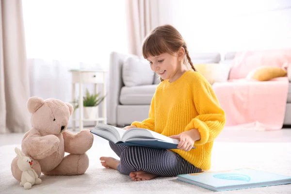 Menina bonito com ursinho de pelúcia leitura livro no chão em casa — Fotografia de Stock