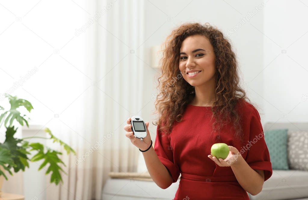Young African-American woman holding digital glucometer and apple at home. Diabetes diet