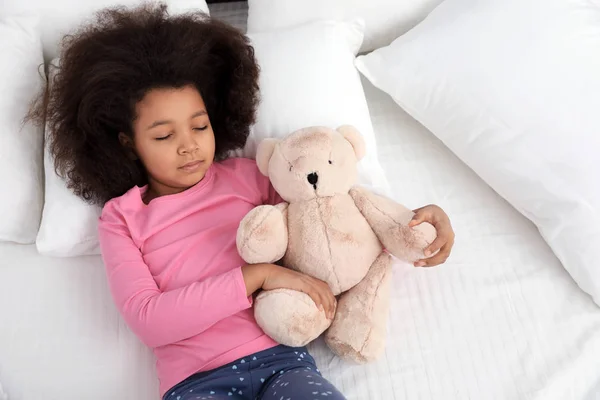 Petite fille afro-américaine mignonne avec ours en peluche dormant au lit, vue sur le dessus — Photo