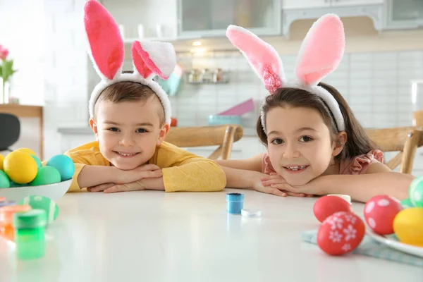 Милые дети с повязками на ушах и раскрашенными пасхальными яйцами, сидящие за столом на кухне — стоковое фото